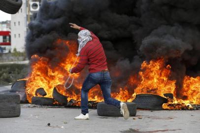 A Palestinian demonstrator holds a sling shot as he runs past burning tyres during clashes with Israeli troops following protests in the West Bank city of Ramallah on December 7, 2017 against a decision by US President Donald Trump to recognise Jerusalem as the capital of Israel.  / AFP PHOTO / ABBAS MOMANI