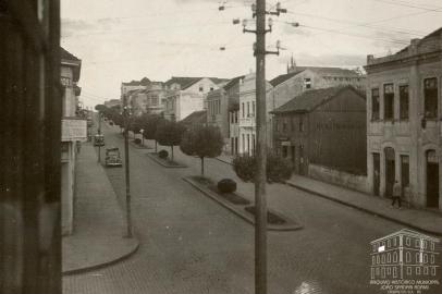 Na foto vê-se o Mercadinho do Povo (casarão de madeira), fundado em 1922 pelo empresário Attilio Mariani. Na imagem, o Mercadinho do Povo em 1947, com a identificação na lateral da casa. À direita, parte do prédio da antiga Farmácia Confiança, na esquina com a Garibaldi. Ao lado, o terreno que, nos anos 1950, viu nascer o Edifício Caberlon. Subindo a Júlio, os sobrados que cederam espaço aos edifícios Prataviera e Jotacê. Sobrevivente do meio do trecho mesmo, só o prédio da antiga sede dos Armarinhos Caxias, atualmente Lojas Uruguai.