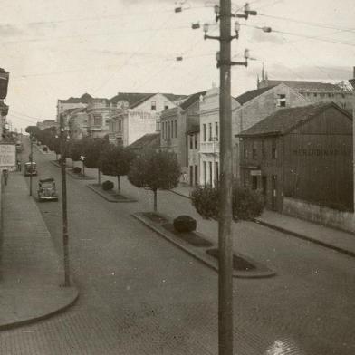 Na foto vê-se o Mercadinho do Povo (casarão de madeira), fundado em 1922 pelo empresário Attilio Mariani. Na imagem, o Mercadinho do Povo em 1947, com a identificação na lateral da casa. À direita, parte do prédio da antiga Farmácia Confiança, na esquina com a Garibaldi. Ao lado, o terreno que, nos anos 1950, viu nascer o Edifício Caberlon. Subindo a Júlio, os sobrados que cederam espaço aos edifícios Prataviera e Jotacê. Sobrevivente do meio do trecho mesmo, só o prédio da antiga sede dos Armarinhos Caxias, atualmente Lojas Uruguai.