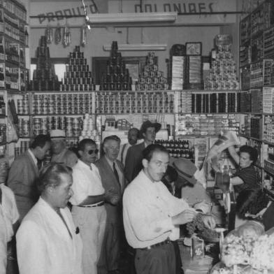 Silla Mariani Santini (á direita, ao fundo, atrás do balcão) e Faustino Ulderico Santini, proprietários da lendária Mercearia caxiense, nos anos 1950. Mercearia foi fundada em 1952, na Avenida Júlio de Castilhos, em frente ao antigo Studio Geremia, entre a Dr. Montaury e a Visconde de Pelotas.