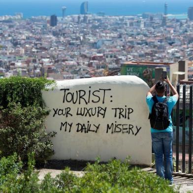 A tourist takes a picture of the citys panorama as he stands next to a wall with a graffito reading Tourist: your luxury trip - my daily misery at Park Guell on August 10, 2017 in Barcelona. Spanish activists in recent weeks have launched initiatives against what they consider a lack of control of mass tourism in cities like Barcelona and Palma de Mallorca, according to media reports. / AFP PHOTO / Josep LAGO