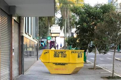 Caçamba para recolher resíduos da construção civil bem no meio de calçada da Avenida Júlio de Castilhos, a 10 metros da Praça Dante Alihgieri, restringe espaço de pedestres e cadeirantes.