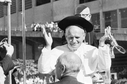  Visita do Papa João Paulo II, em Porto Alegre, no sábado, 5 de julho de 1980.O Sumo Pontífice é recebido diante do Gigantinho por 200 cavalarianos pilchados. Grupos folclóricos apresentam três danças típicas: o pezinho, o balaio e a cana verde. O Papa assiste à homenagem até o fim. O folclorista Paixão Côrtes oferece uma cuia de chimarrão ao visitante. Desconfiado, pergunta do que se trata.- É um sinal de amizade - esclarece dom Vicente Scherer.Enquanto o Papa sorve o chimarrão, um chapéu de gaúcho é colocado sobre sua cabeça.