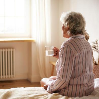 Rear view of senior sixty year old woman with gray hair holding mug washing down sleeping pill, suffering from insomnia. Elderly retired female taking medicine with water, sitting in bedroomPORTO ALEGRE, RS, BRASIL, 22/10/2019- Senhora tomando remédio. (Foto: shurkin_son / stock.adobe.com)Fonte: 284095310