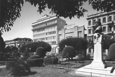 Praça Dante Alighieri (antiga Praça Rui Barbosa) em meados dos anos 1950, quando as árvores eram podadas segundo os pincípios da topiaria (esculturas vivas). Ao fundo, os prédios do Banrisul, Cine Guarany, Hotel Menegotto e Clube Juvenil. À direita, o busto de Dante Alighieri.