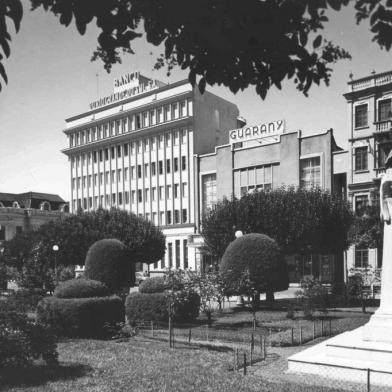 Praça Dante Alighieri (antiga Praça Rui Barbosa) em meados dos anos 1950, quando as árvores eram podadas segundo os pincípios da topiaria (esculturas vivas). Ao fundo, os prédios do Banrisul, Cine Guarany, Hotel Menegotto e Clube Juvenil. À direita, o busto de Dante Alighieri.