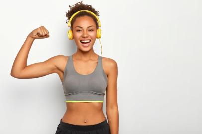 Horizontal view of dark skinned woman in good mood, raises arm with muscles, has strong body, dressed in gym outfit, listens audio via modern headphones, poses indoor. Fitness and music conceptVista horizontal da mulher de pele escura de bom humor, levanta o braço com músculos, tem corpo forte, vestido com roupa de ginástica, escuta áudio através de fones de ouvido modernos, poses indoor. Fitness e conceito de músicaFonte: 294064530<!-- NICAID(14316643) -->