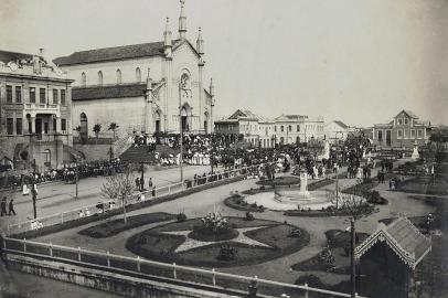 Quase não há fotos do centro de Caxias durante o surto de gripe espanhola, entre finais de 1918 e 1919, tampouco registros das medidas sanitárias junto aos vagões da Estação Férrea. O registro acima, do início da década de 1920, traz o cenário da Praça Dante Alighieri pós-epidemia, uma concentração de pessoas impensável dois anos antes, quando a cidade recolheu-se em casa. Trata-se de uma cerimônia religiosa captada a partir da Rua Marquês do Herval, com o recém-construído prédio do Bispado, a Catedral Diocesana, o casarão que abrigou a loja A Economia do Povo (embrião da Casa Magnabosco, de Raymundo Magnabosco) e o casarão de alvenaria da família Serafini (onde viveu a jovem Flora Serafini, filha do fotógrafo Giovanni Battista Serafini e esposa de Raymundo). Na praça ainda cercada por muros, o busto de Dante Alighieri, surgido em 1914 e envolto por grades (à frente), e o de Júlio Prates de Castilhos (ao fundo).<!-- NICAID(14468745) -->