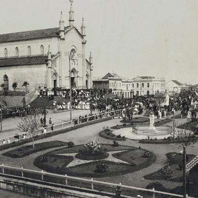 Quase não há fotos do centro de Caxias durante o surto de gripe espanhola, entre finais de 1918 e 1919, tampouco registros das medidas sanitárias junto aos vagões da Estação Férrea. O registro acima, do início da década de 1920, traz o cenário da Praça Dante Alighieri pós-epidemia, uma concentração de pessoas impensável dois anos antes, quando a cidade recolheu-se em casa. Trata-se de uma cerimônia religiosa captada a partir da Rua Marquês do Herval, com o recém-construído prédio do Bispado, a Catedral Diocesana, o casarão que abrigou a loja A Economia do Povo (embrião da Casa Magnabosco, de Raymundo Magnabosco) e o casarão de alvenaria da família Serafini (onde viveu a jovem Flora Serafini, filha do fotógrafo Giovanni Battista Serafini e esposa de Raymundo). Na praça ainda cercada por muros, o busto de Dante Alighieri, surgido em 1914 e envolto por grades (à frente), e o de Júlio Prates de Castilhos (ao fundo).<!-- NICAID(14468745) -->