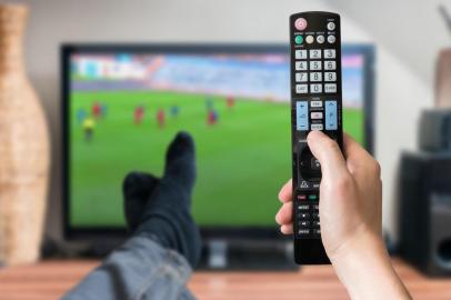  Man is relaxing with legs on table and is watching football match on tv and holds remote controller.Fonte: 113833428<!-- NICAID(14535162) -->