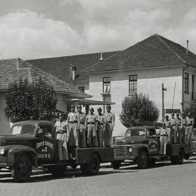 O comandante Frederico Bergmann (à esquerda) com os bombeiros e os clássicos modelos Ford na Rua Sinimbu, próximo à Coronel Flores, onde ficava a antiga sede. O prédio à direita abrigava, no piso superior, a Congregação das Irmãs Carlistas (Colégio São Carlos). No térreo, o alojamento da Brigada Militar e dos bombeiros.  A partir da esquerda vê-se o então comandante Frederico Bergmann, o sargento Oscar de Azevedo, Nei Andrade, Artur Mello, Joecy da Silva, Candido Abreu e João Maria Machado, entre outros não identificados. Os veículos Ford 1946/1948 eram modelos auto-bomba-tanque.<!-- NICAID(14537762) -->