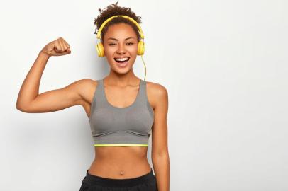 Horizontal view of dark skinned woman in good mood, raises arm with muscles, has strong body, dressed in gym outfit, listens audio via modern headphones, poses indoor. Fitness and music conceptVista horizontal da mulher de pele escura de bom humor, levanta o braço com músculos, tem corpo forte, vestido com roupa de ginástica, escuta áudio através de fones de ouvido modernos, poses indoor. Fitness e conceito de músicaFonte: 294064530<!-- NICAID(14316643) -->