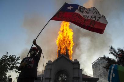  A demonstrator flutters a Chilean flag outside the burning church of Asuncion, set on fire by protesters, on the commemoration of the first anniversary of the social uprising in Chile, in Santiago, on October 18, 2020, as the country prepares for a landmark referendum. - Two churches were torched as tens of thousands of demonstrators gathered Sunday in a central Santiago square to mark the anniversary of a protest movement that broke out last year demanding greater equality in Chile. The demonstration comes just a week before Chileans vote in a referendum on whether to replace the dictatorship-era constitution -- one of the key demands when the protest movement began on October 18, 2019. (Photo by MARTIN BERNETTI / AFP)Editoria: POLLocal: SantiagoIndexador: MARTIN BERNETTISecao: crisisFonte: AFPFotógrafo: STF<!-- NICAID(14619968) -->