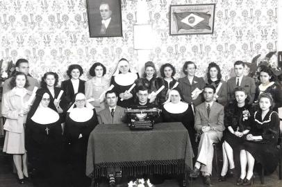 Caxias, dezembro de 1943: a foto oficial dos formandos do curso de datilografia no Colégio São Carlos, com a jovem Ignez Boff (de roupa clara, abaixo do quadro de Vargas) e o pároco Eugênio Giordani (sentado ao centro).<!-- NICAID(14904845) -->