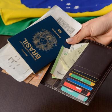 Hand holding Brazilian passport over table with Brazilian flag in the background.*A PEDIDO DE GABRIELA PERUFO* Hand holding Brazilian passport over table with Brazilian flag in the background. - Foto: Cacio Murilo/stock.adobe.comIndexador: Cacio Murilo de VasconcelosFonte: 523946492Fotógrafo: FotÃ³grafo<!-- NICAID(15195610) -->